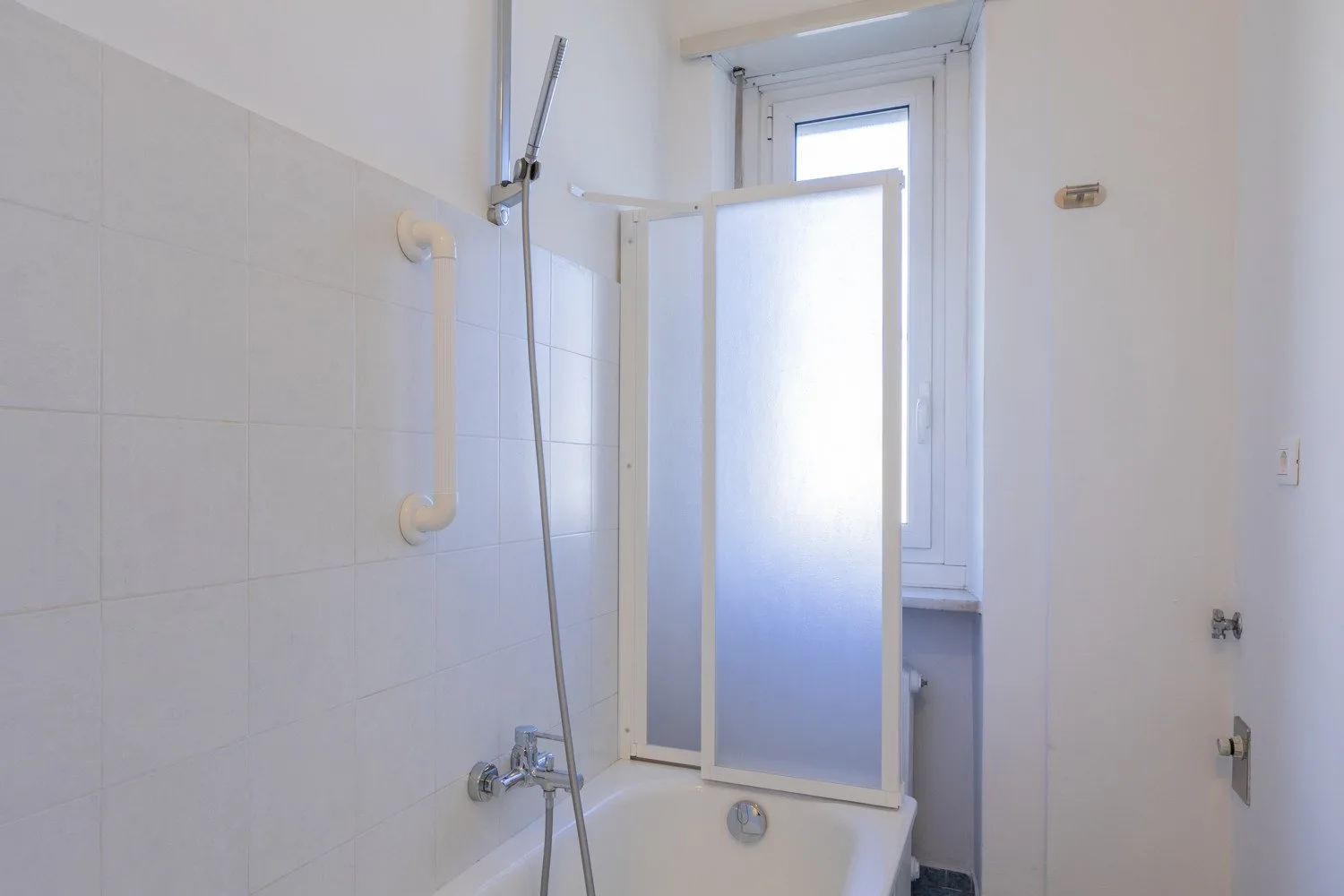 A tiled bathroom with a bathtub, handheld shower, and frosted glass screen.