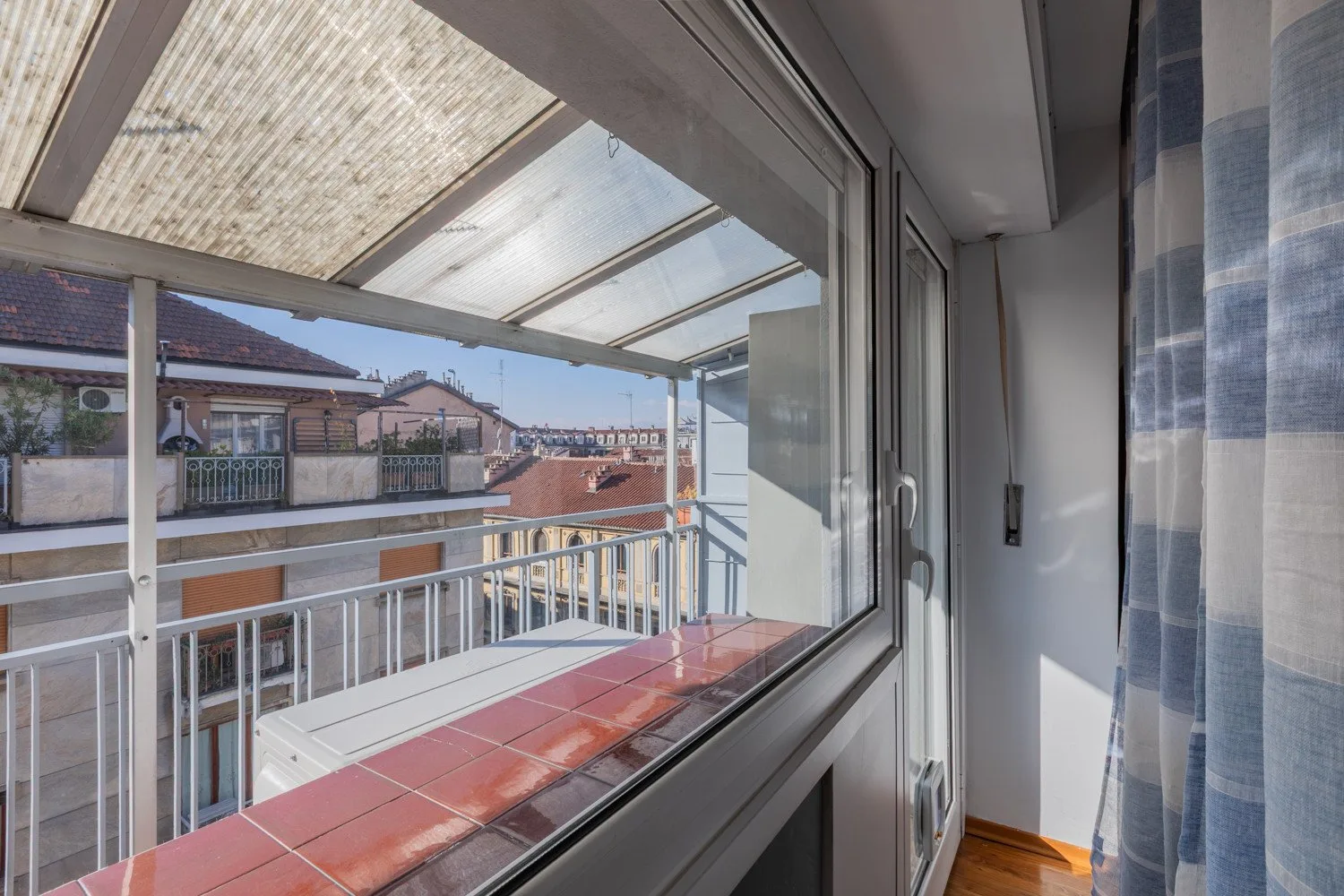 A bright enclosed balcony with glass panels overlooks nearby rooftops.
