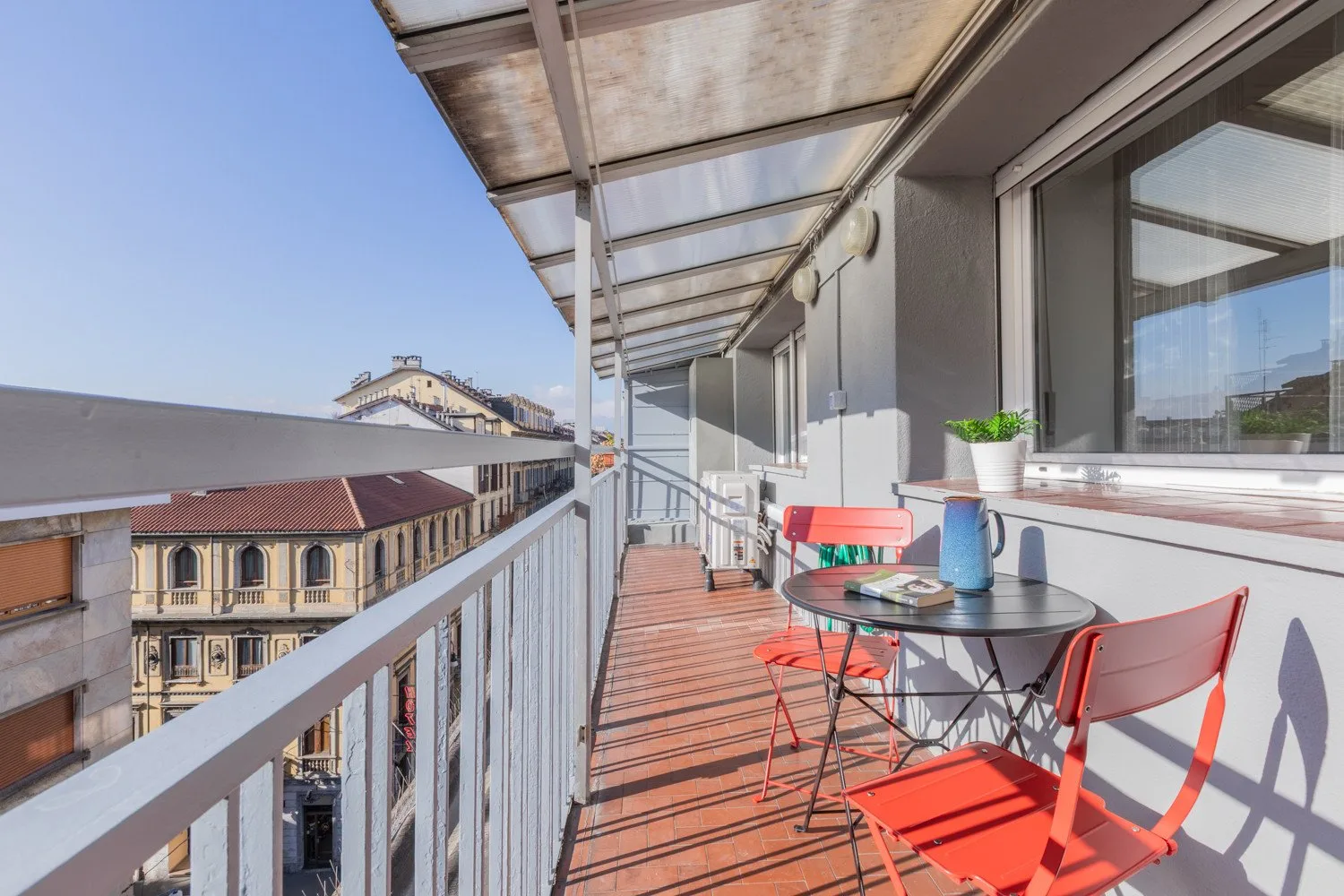 A narrow balcony with a small table and red seating overlooks neighboring buildings.