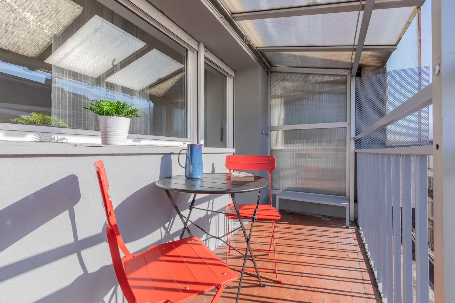 A sunlit enclosed balcony has a small table, red seating, and potted plants.