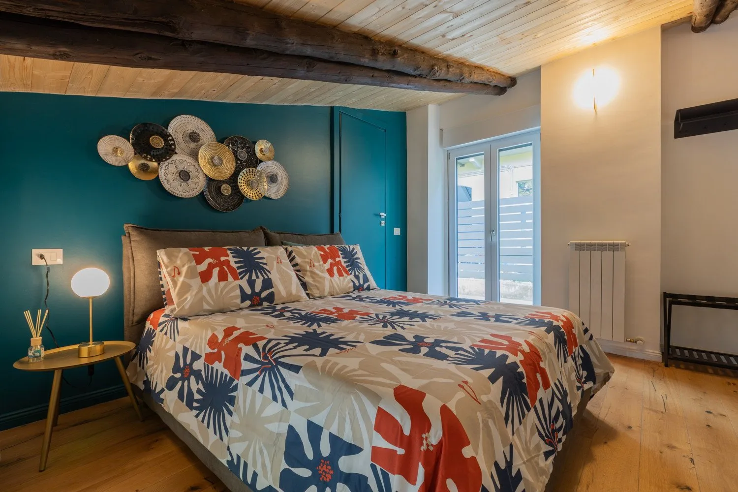 A bedroom with a colorful patterned bed, teal wall, and large windows.