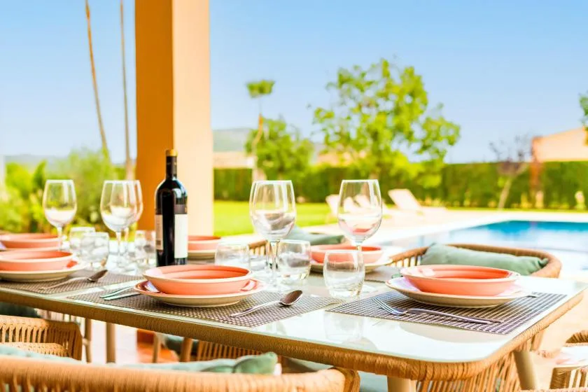 Una mesa de comedor al aire libre está puesta con platos y vasos junto a una piscina.