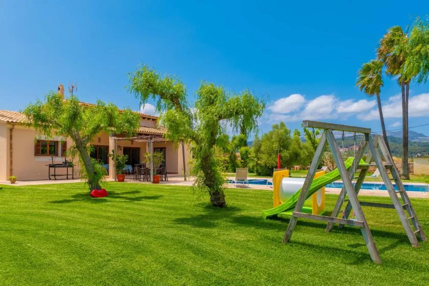 Un jardín con una estructura de juegos, una piscina y una casa al fondo.