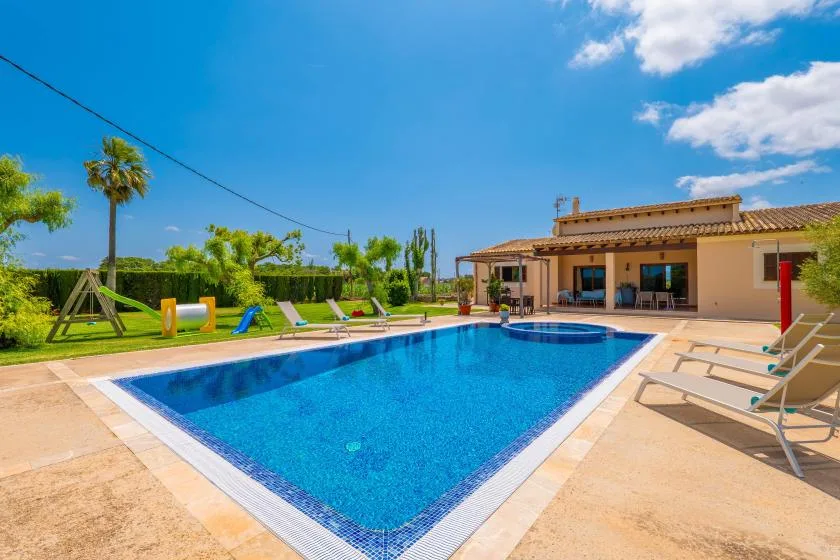 Piscina exterior junto a una casa de una planta, con tumbonas y jardín.