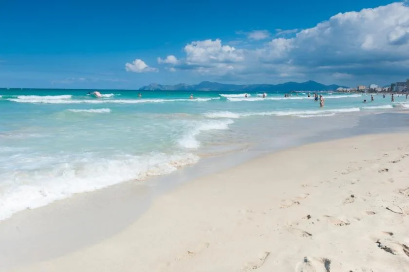 Una playa de arena se encuentra con olas turquesas bajo un cielo azul con nubes.