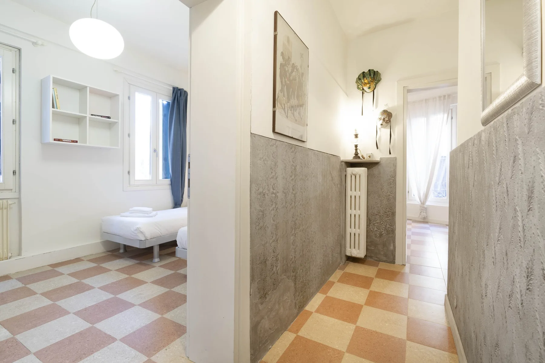 Un pasillo luminoso con suelo de baldosas y una cama visible en la habitación contigua.