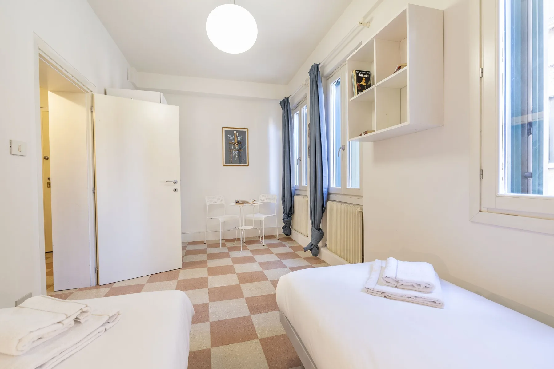 Un dormitorio luminoso con camas individuales, una mesa pequeña y ventanas amplias.