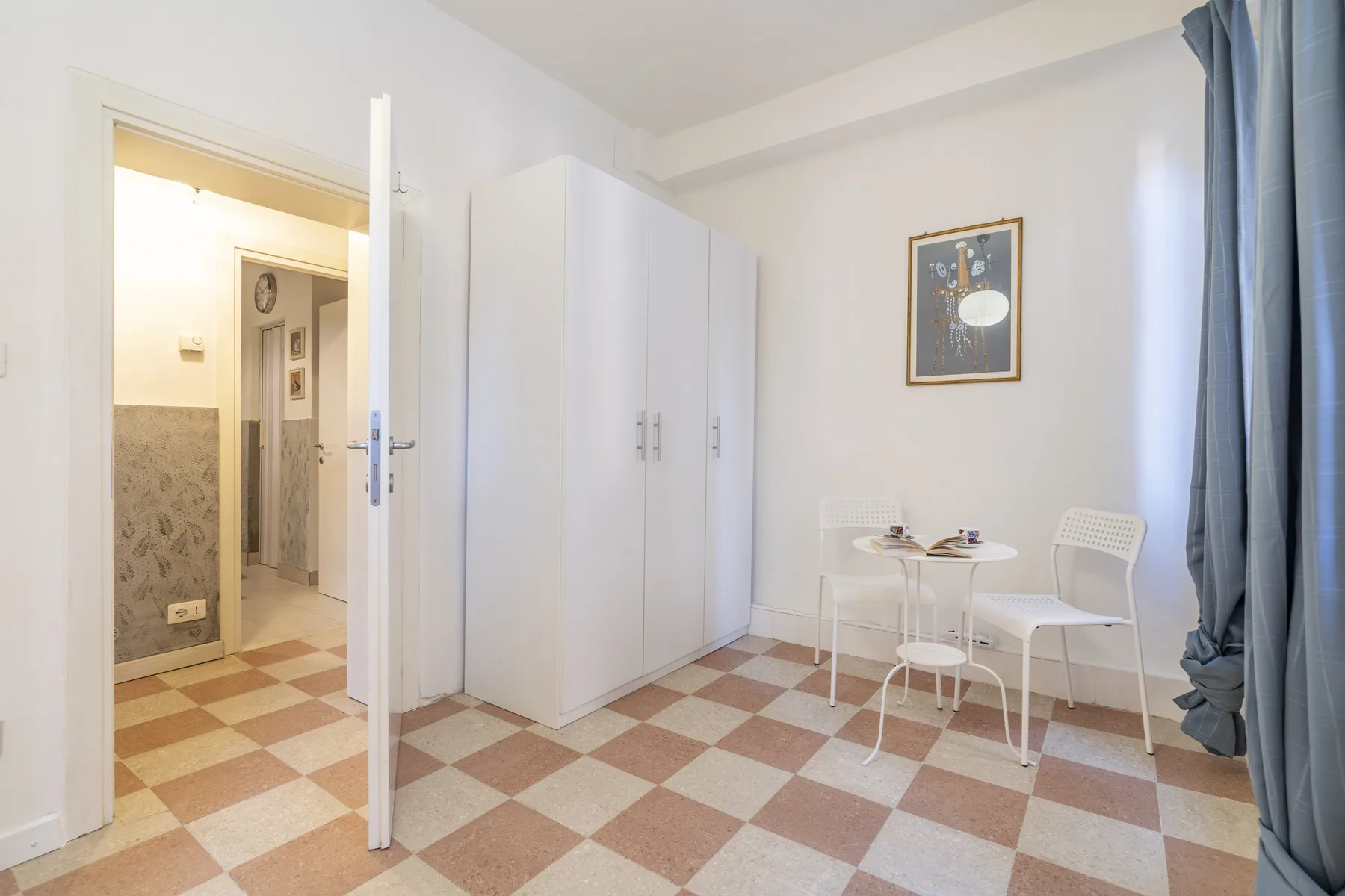 Una habitación luminosa con armario, una mesa pequeña y sillas junto a una ventana con cortinas.
