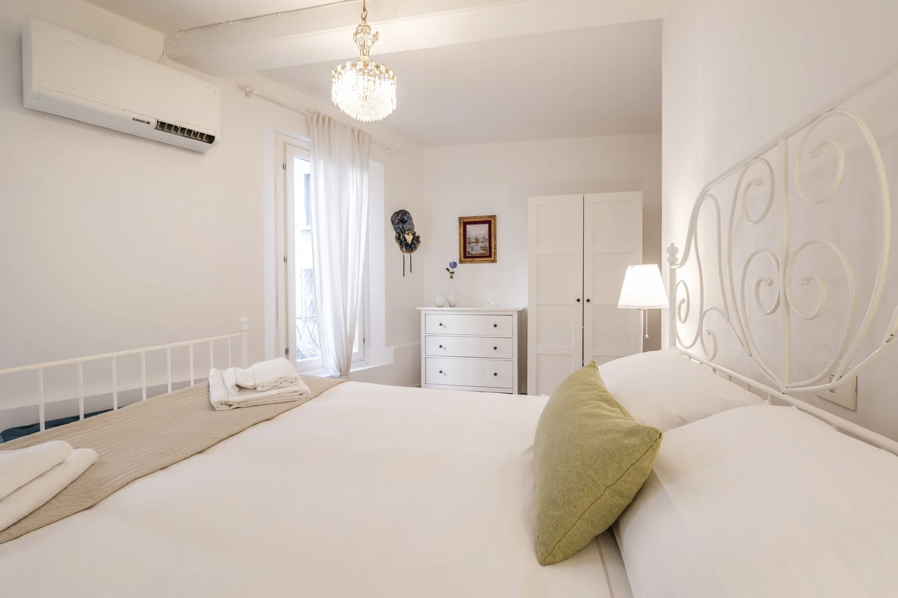 Un dormitorio luminoso con una cama grande, una cómoda y una ventana con cortinas translúcidas.