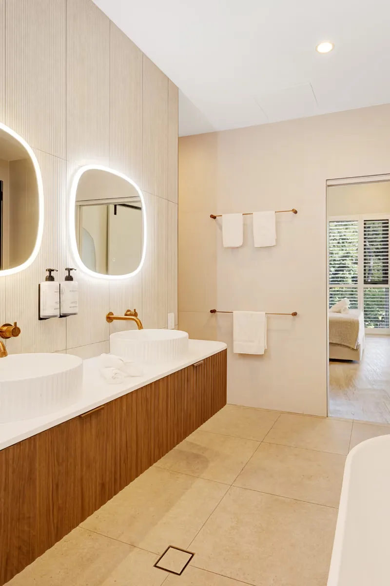 Baño de huéspedes con dos lavabos, espejos retroiluminados y grifería de latón.