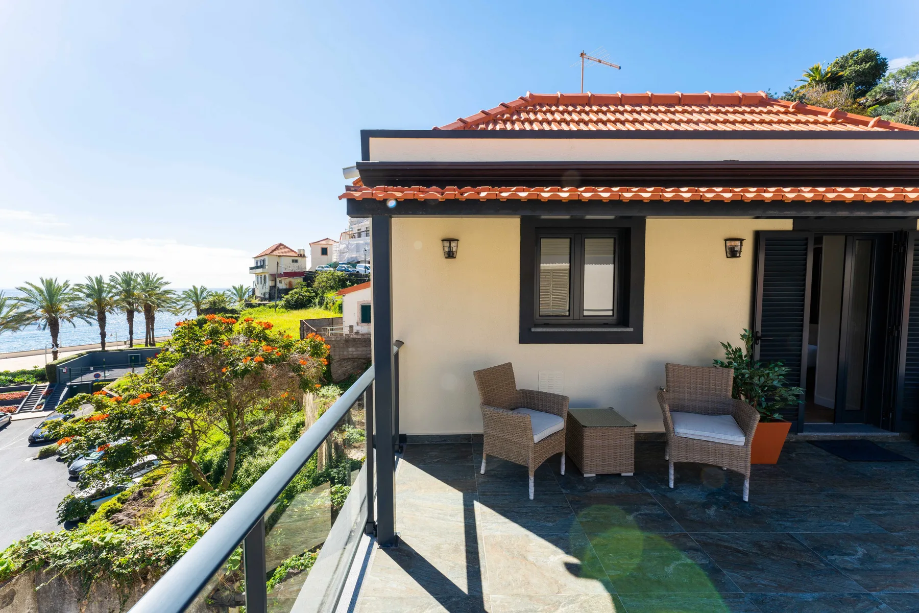 Una terraza soleada tiene asientos al aire libre junto a una barandilla de vidrio con vistas a la costa.