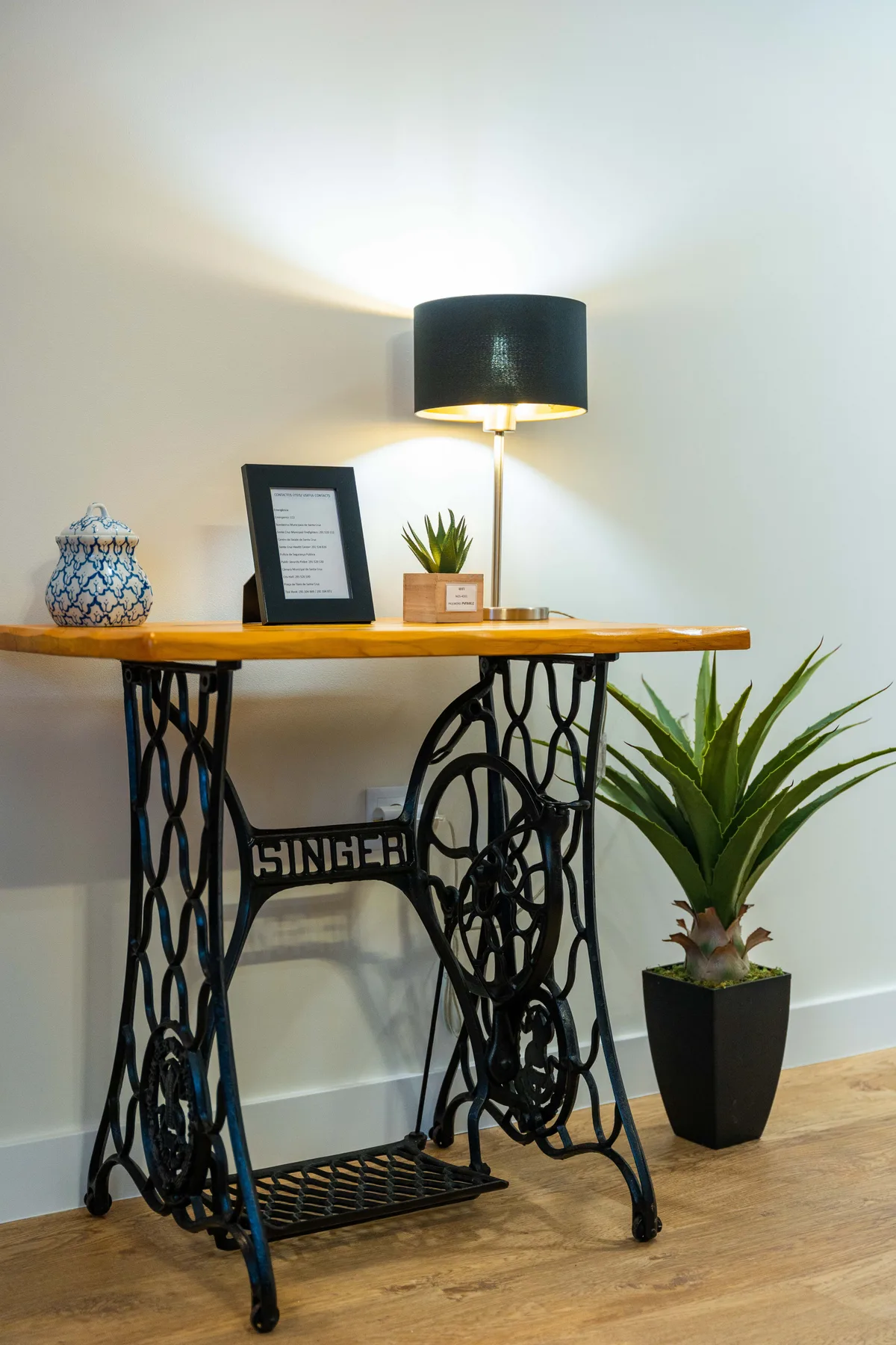 Una mesa consola decorativa con una lámpara y plantas en macetas está junto a una pared clara.