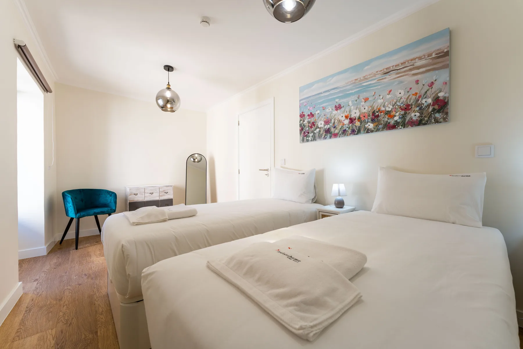 Un dormitorio luminoso con dos camas, una silla azul y un cuadro floral.