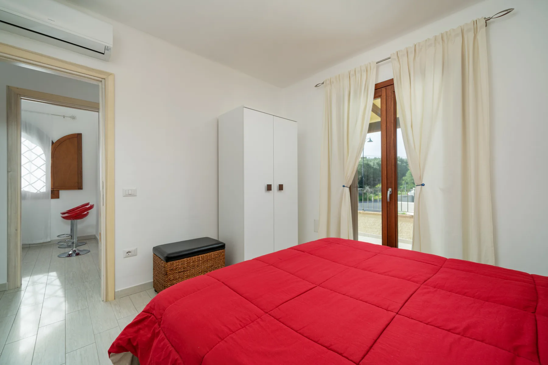 Un dormitorio con una colcha roja, un armario y cortinas junto a una puerta de vidrio.