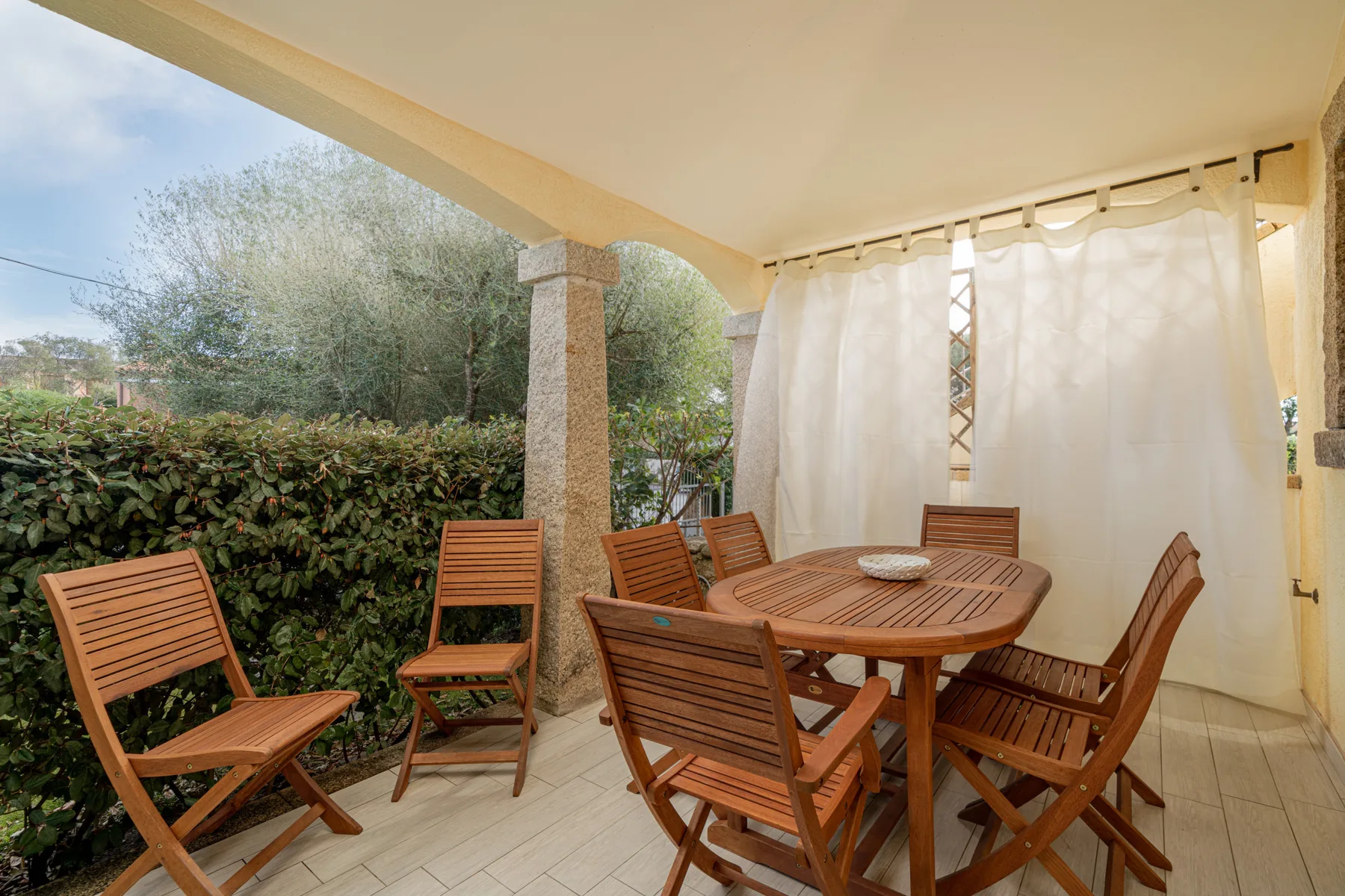 Un patio cubierto con una mesa de madera y asientos junto a un seto frondoso.