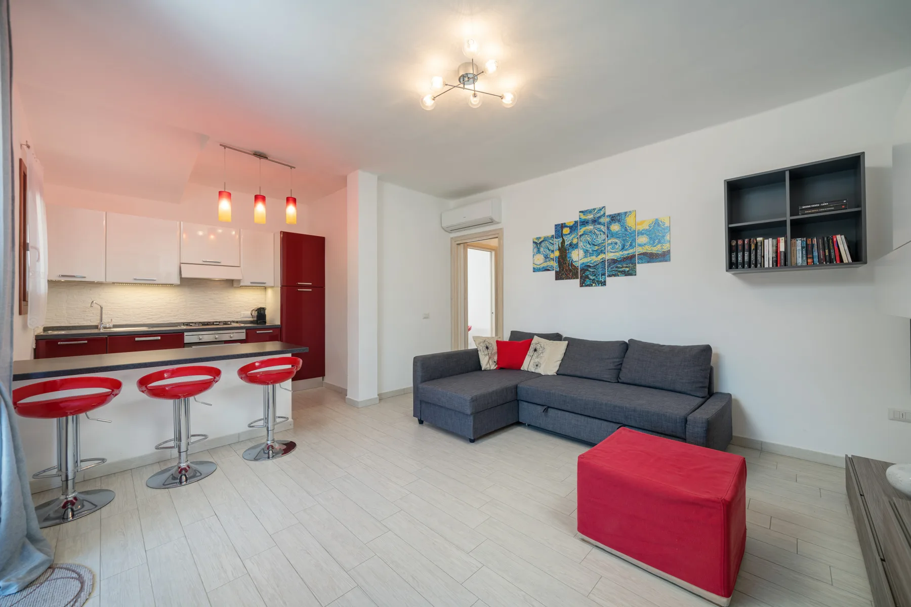 Sala de estar y cocina con asientos grises y un otomano rojo.