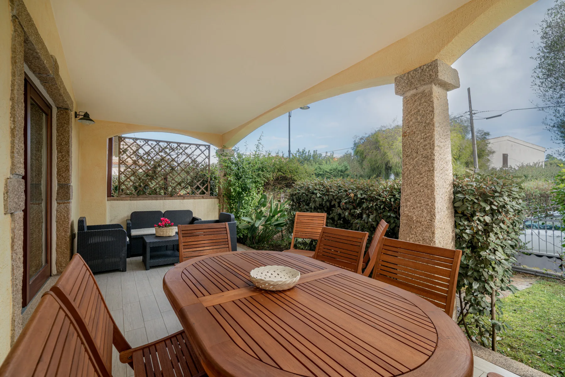 Un patio cubierto con una mesa de comedor de madera y asientos junto a setos frondosos.