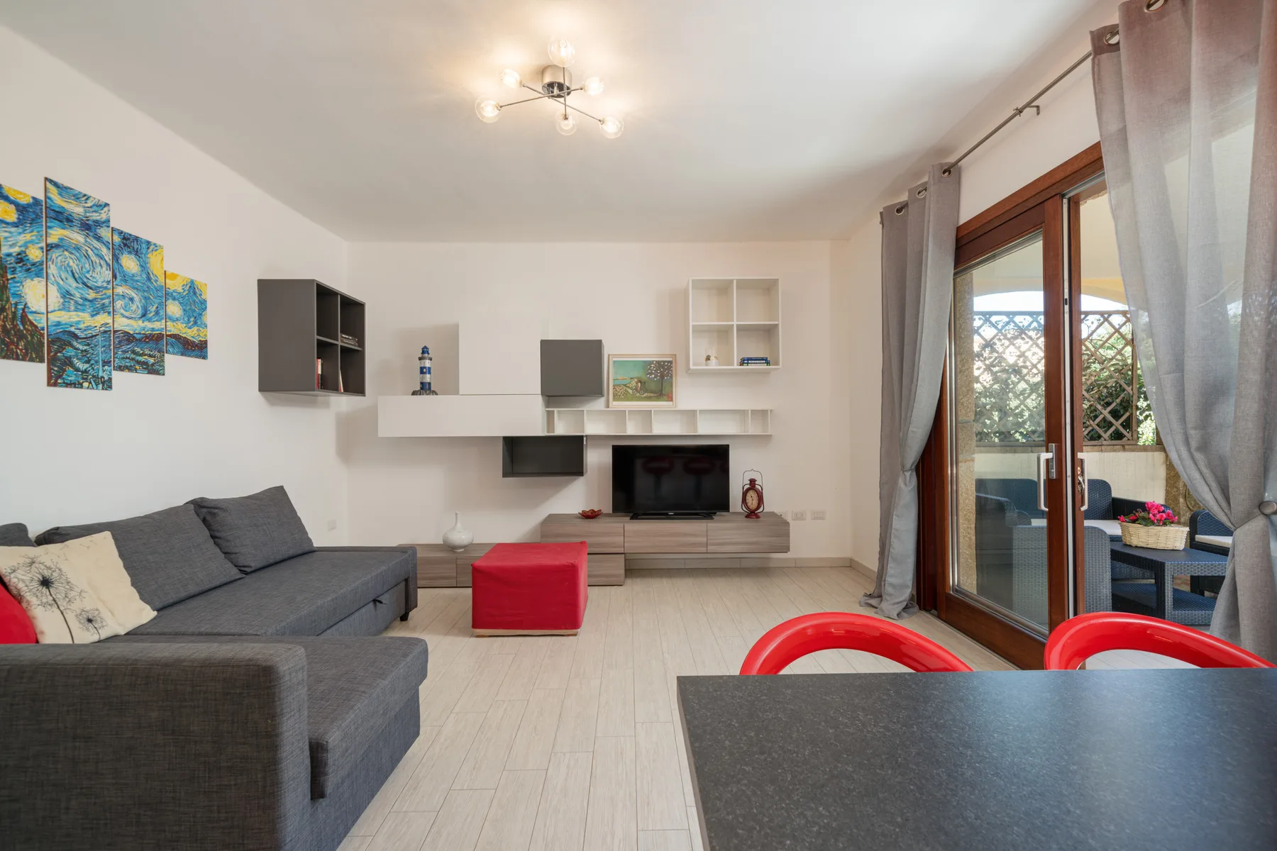 Una sala de estar con sofá seccional gris, estantes de pared y puertas de vidrio hacia un balcón.