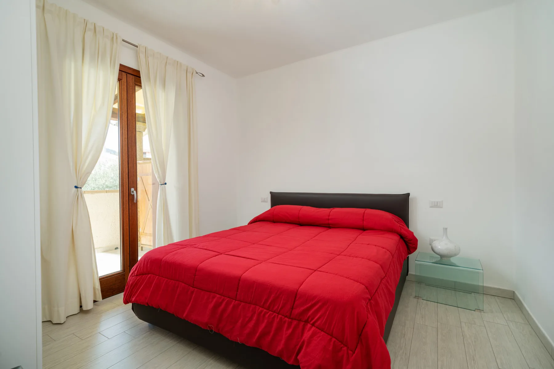 Un dormitorio con una cama roja junto a una puerta de vidrio y cortinas claras.