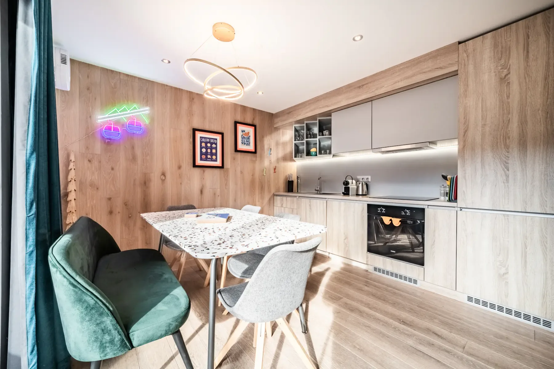 Modern kitchen and dining area with light wood cabinets, terrazzo table, assorted chairs, built-in oven, and neon wall art. Circular ceiling light fixture overhead.