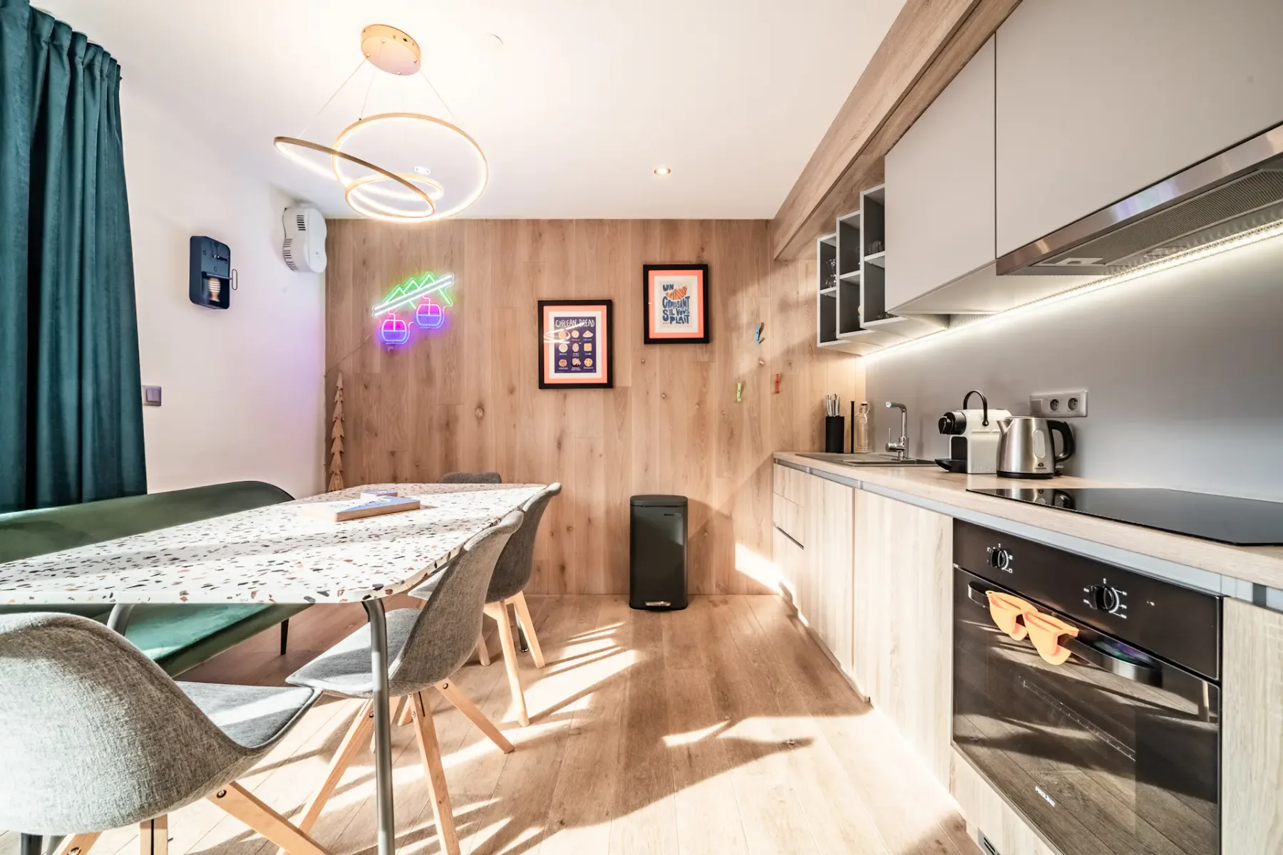 Modern kitchen with wooden cabinets, dining table, gray chairs, wall art, neon lights, and sleek appliances. Bright and airy with natural light.