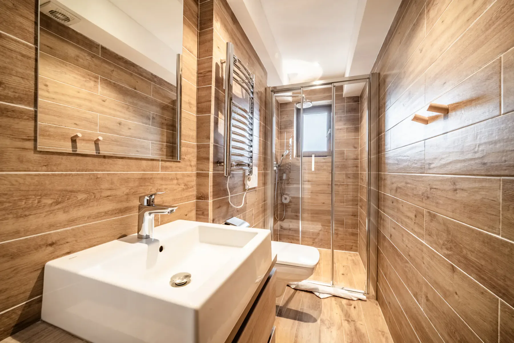 Modern bathroom with wood-patterned walls and flooring, featuring a square sink, wall-mounted toilet, glass shower enclosure, and a small window.