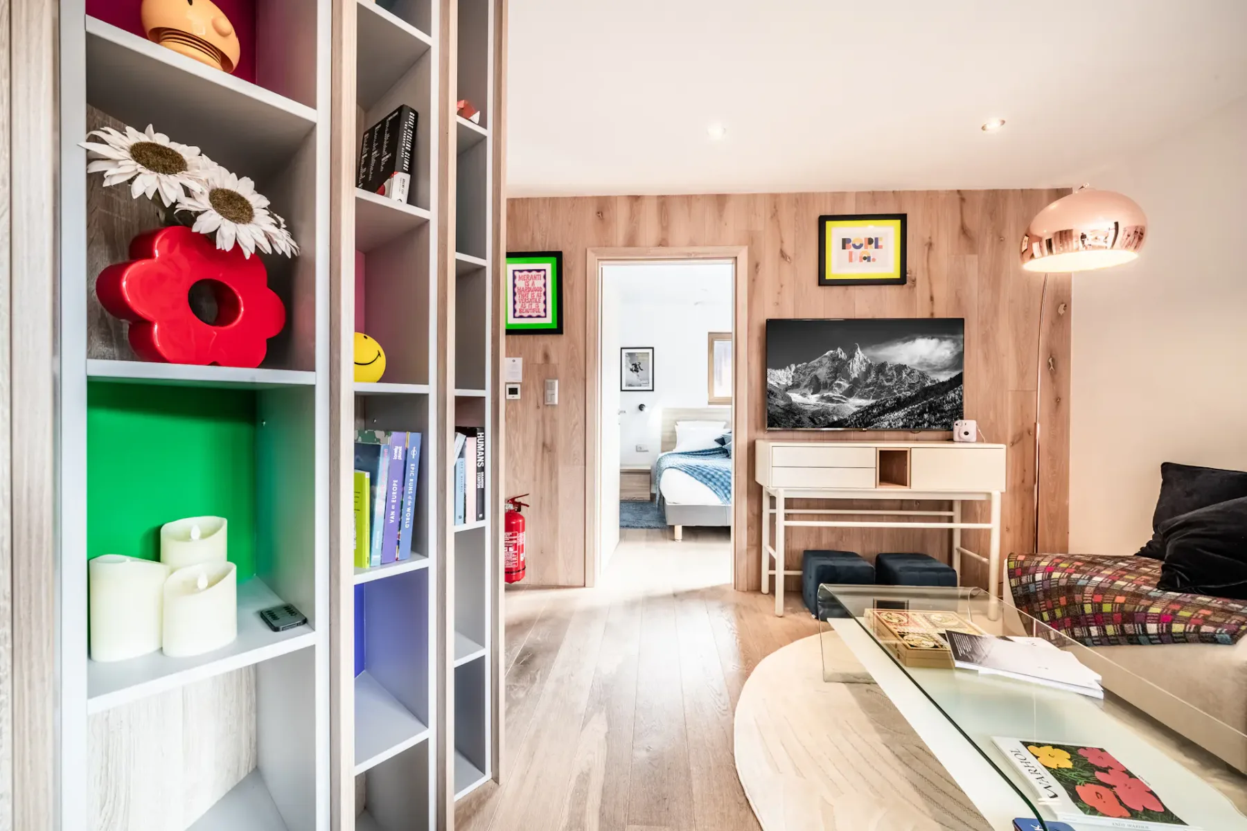 Modern living room with wooden walls, open shelving, framed art, a TV on a small table, glass coffee table, and a view into a bedroom.