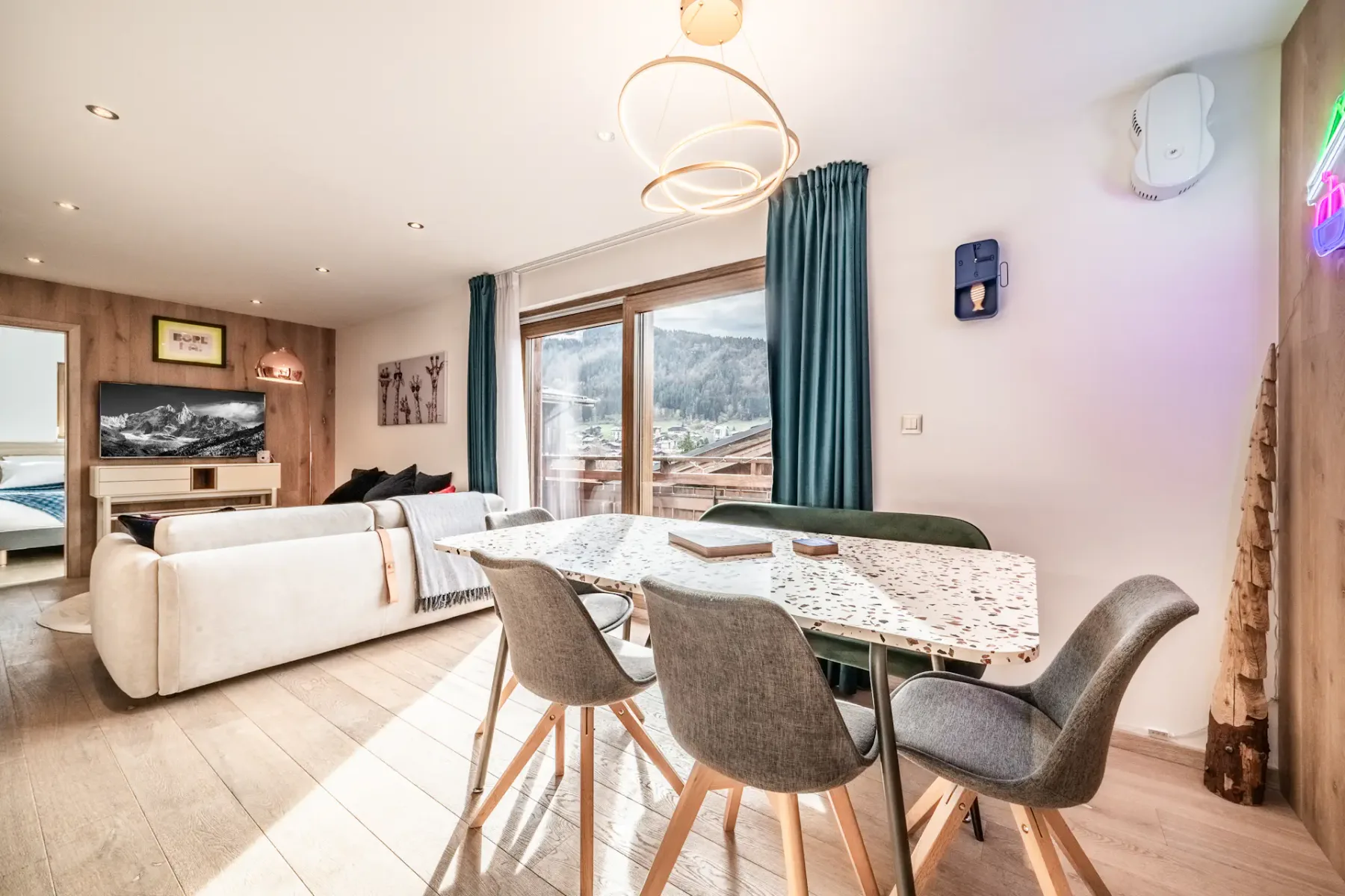 Bright modern living and dining area with a terrazzo table, grey chairs, light wood floors, sofa, wall art, large windows, and a view of mountains outside.