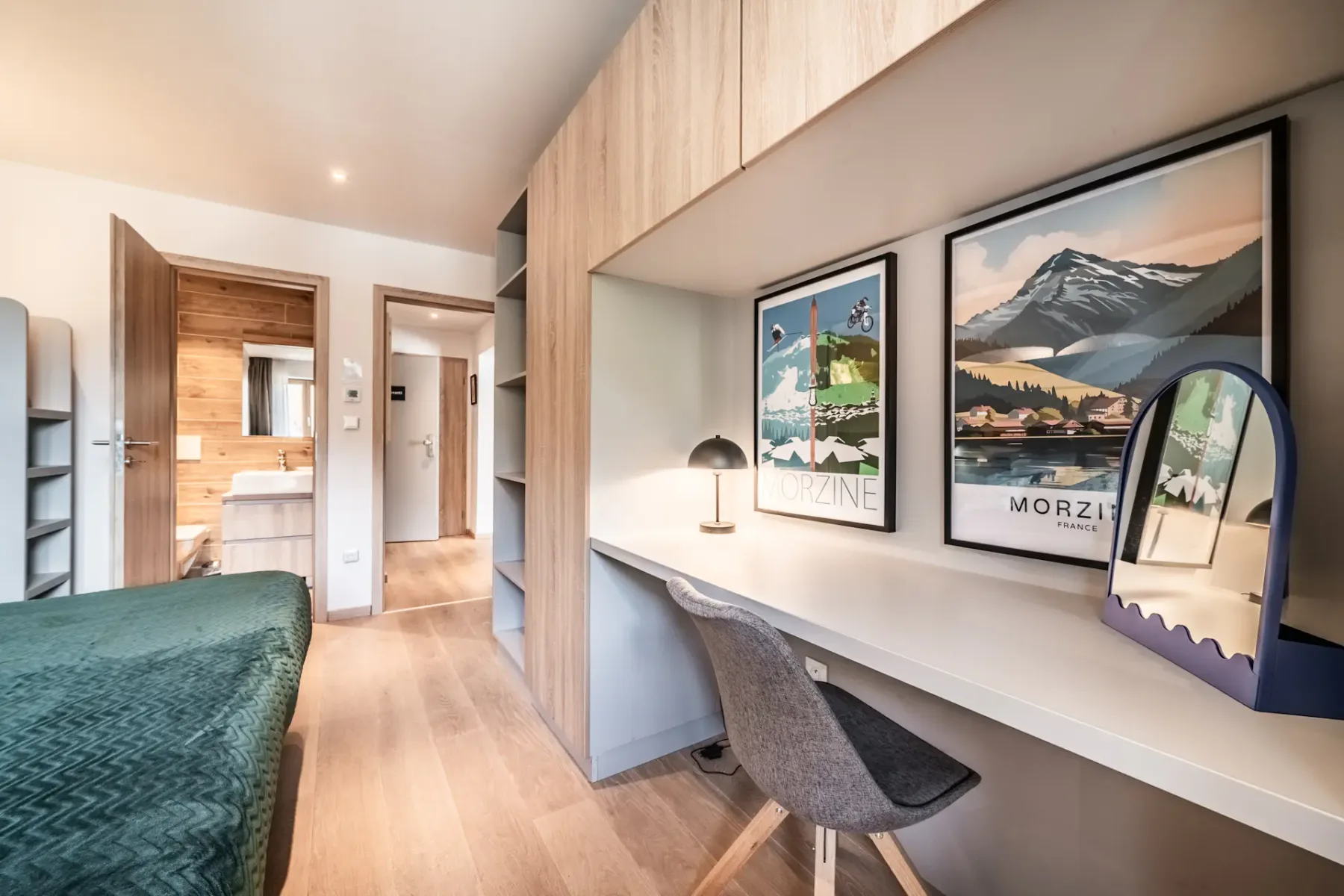 Modern bedroom with a built-in desk, chair, lamp, and vanity mirror. Two travel posters hang above the desk; an open door reveals a bathroom with wood accents.