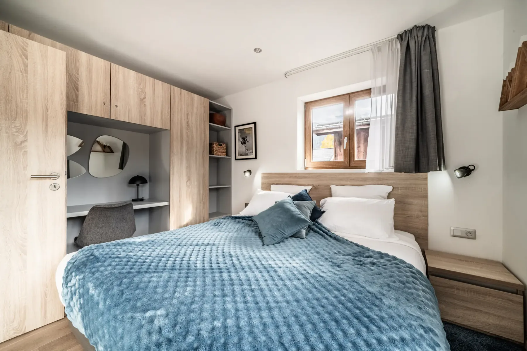 Modern bedroom with a wooden bed, blue textured blanket, built-in wardrobe, small desk with mirror, and window with sheer and blackout curtains.