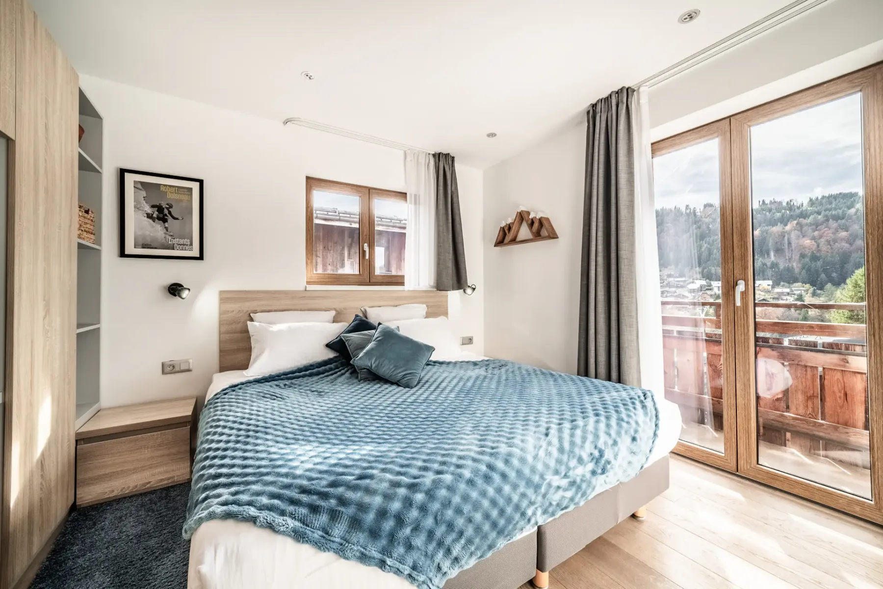 Bright bedroom with a double bed, blue blanket, wall art, wooden wardrobe, and glass doors opening to a balcony with a scenic mountain view.