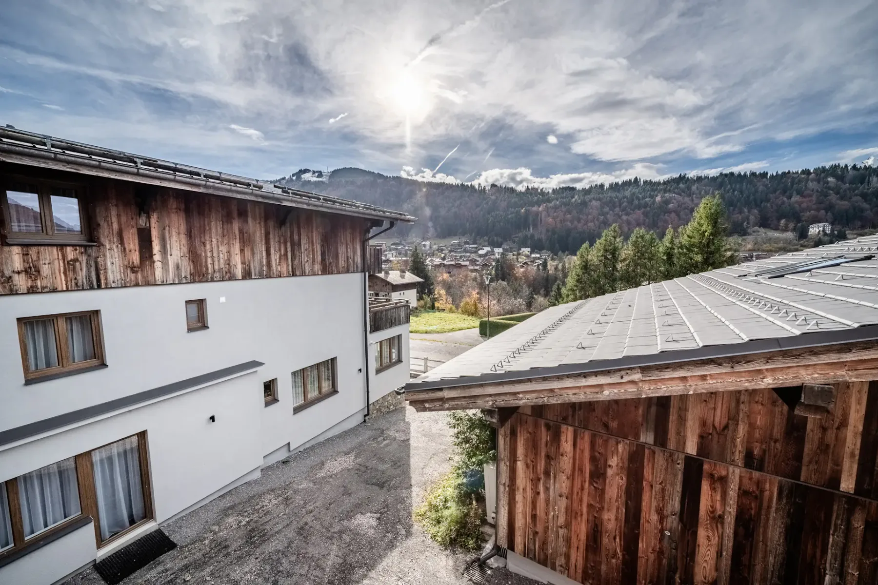 Wooden cabin-style buildings with a mountain landscape and forest in the background under a partly cloudy sky with the sun shining.
