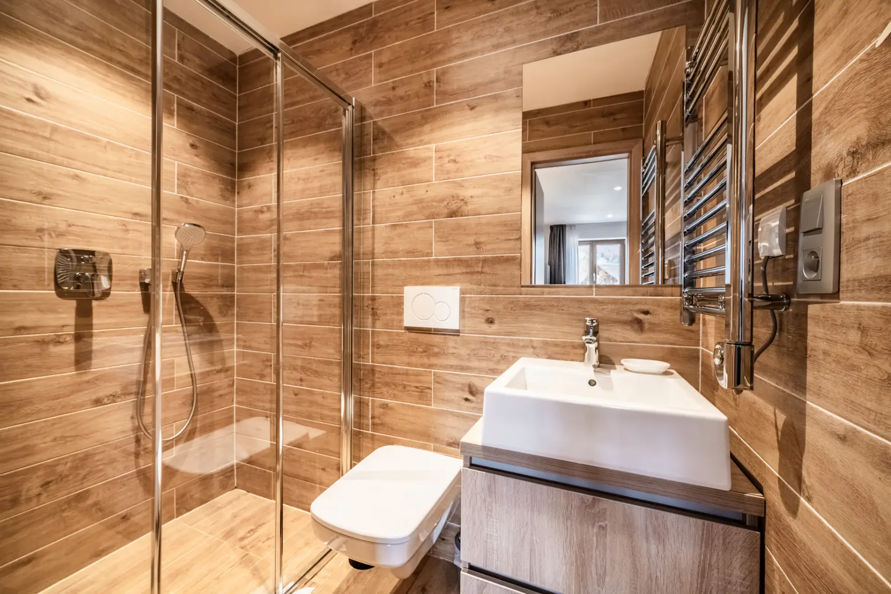 Modern bathroom with wood-look tile walls, a glass-enclosed shower, wall-mounted toilet, rectangular sink, large mirror, and stainless steel towel rack.