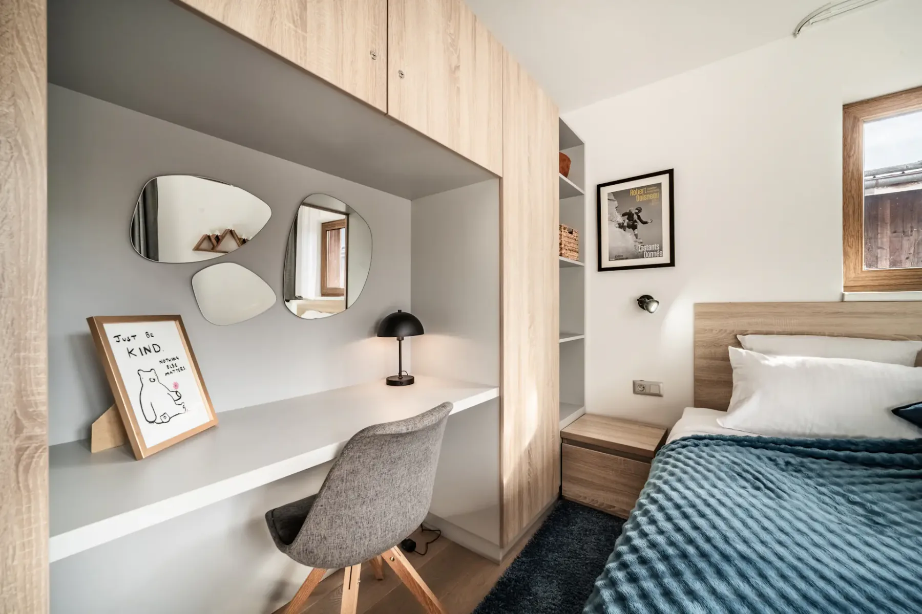 A modern bedroom with a wooden bed, a gray chair at a built-in desk, three irregular mirrors, a framed drawing, shelves, and a window letting in natural light.