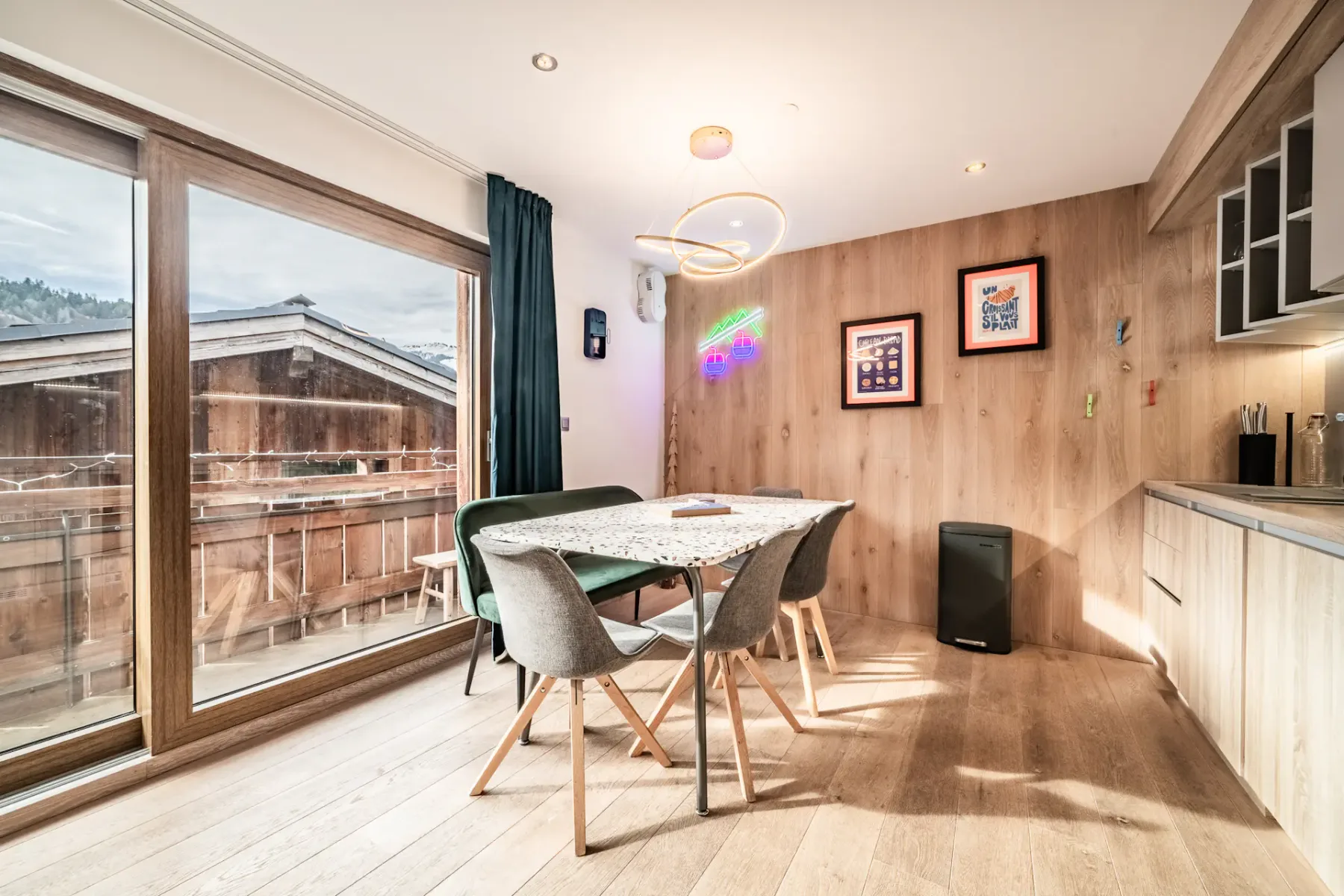 Bright dining area with a table and four chairs, wood-paneled walls, modern lighting, wall art, large windows, and a view of wooden buildings outside.