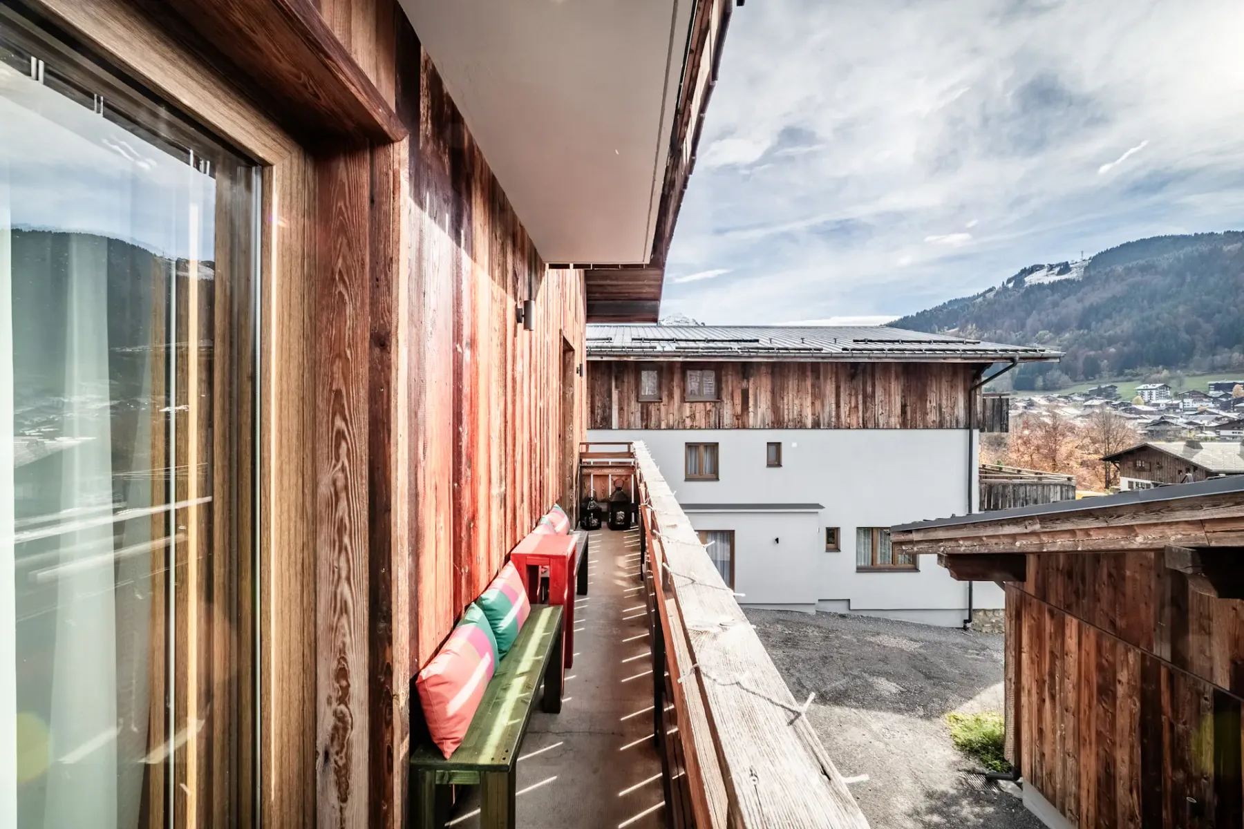 A wooden balcony with colorful cushions on a bench overlooks a courtyard with rustic buildings and mountains in the background under a partly cloudy sky.