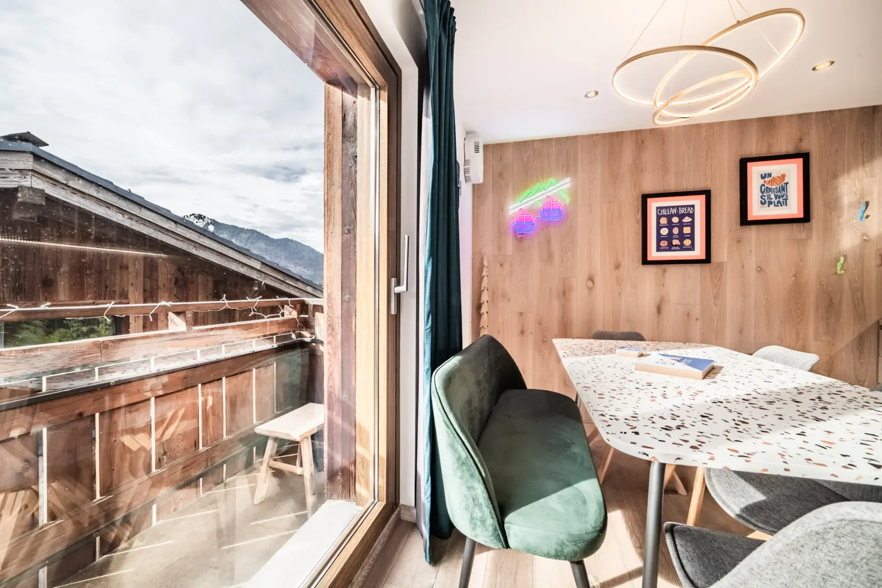 Modern dining area with terrazzo table, green chairs, and mountain view through large glass door; wooden walls feature framed art and neon lights.