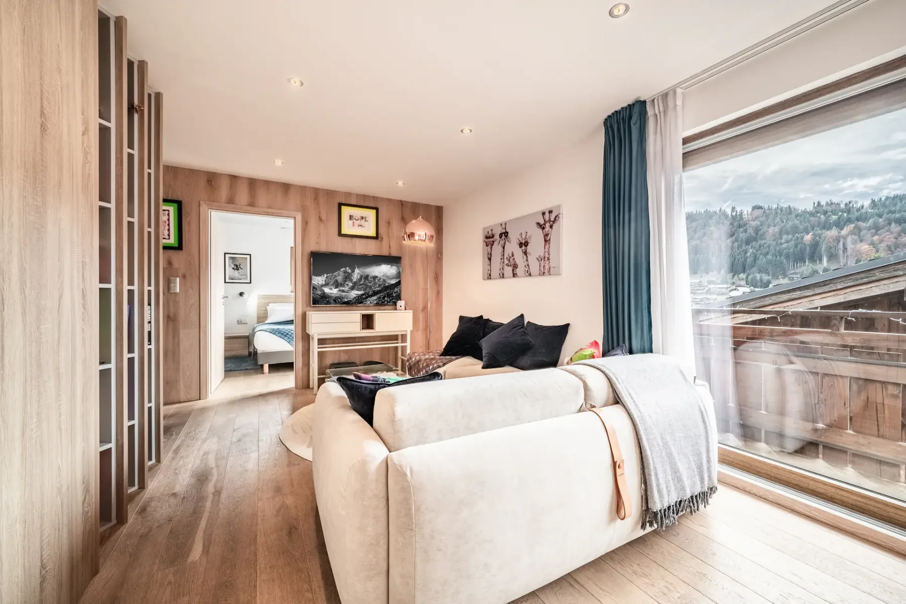 Modern living room with light wood flooring, a white sofa, wall art, and large window overlooking a forested hillside; bedroom visible through open doorway.