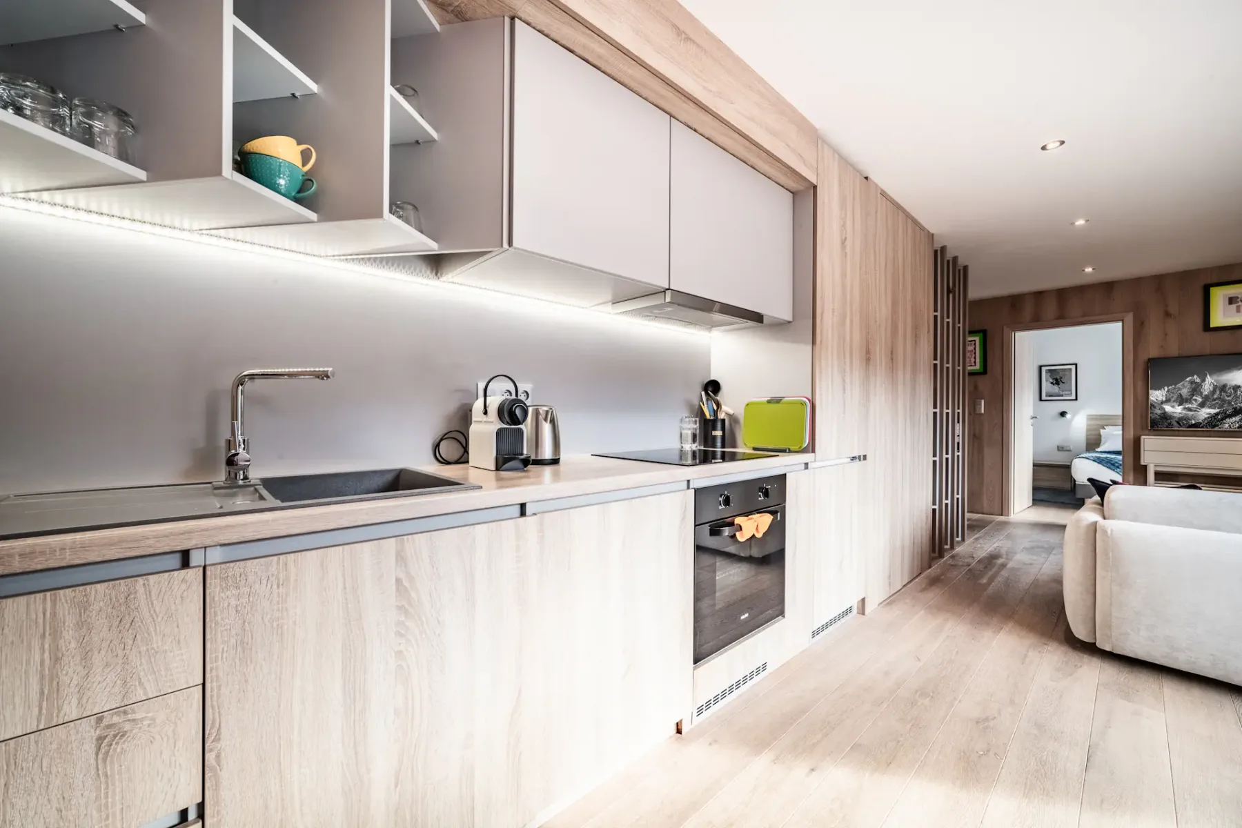 Modern kitchen with light wood cabinets, built-in oven, sink, open shelves with cups, and under-cabinet lighting, extending into a living area and bedroom in the background.
