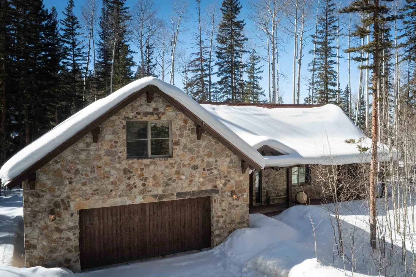 A stone house with a snow-covered roof stands among tall trees.