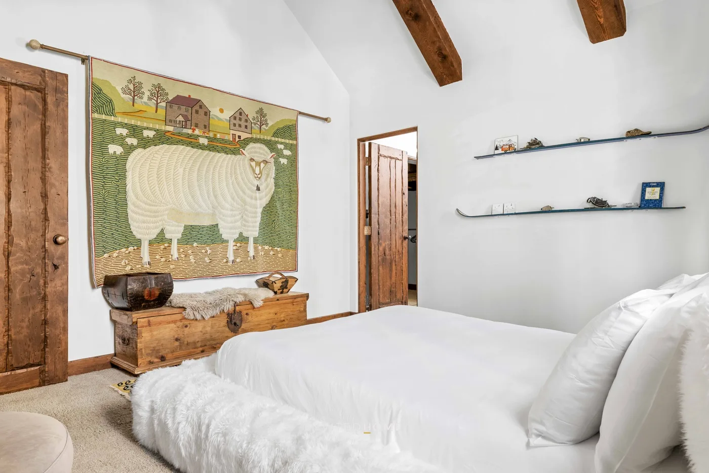 A bedroom with white bedding, wooden ceiling beams, and a sheep wall tapestry.