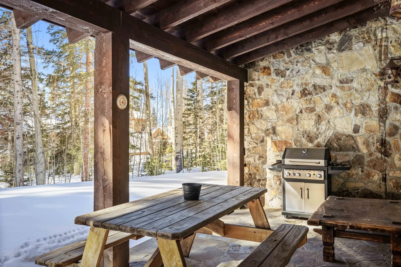 A covered outdoor dining area with a wooden picnic table beside a stone wall.