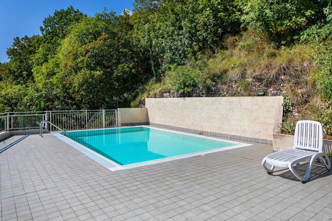 Una piscina al aire libre junto a un patio pavimentado y una ladera arbolada.