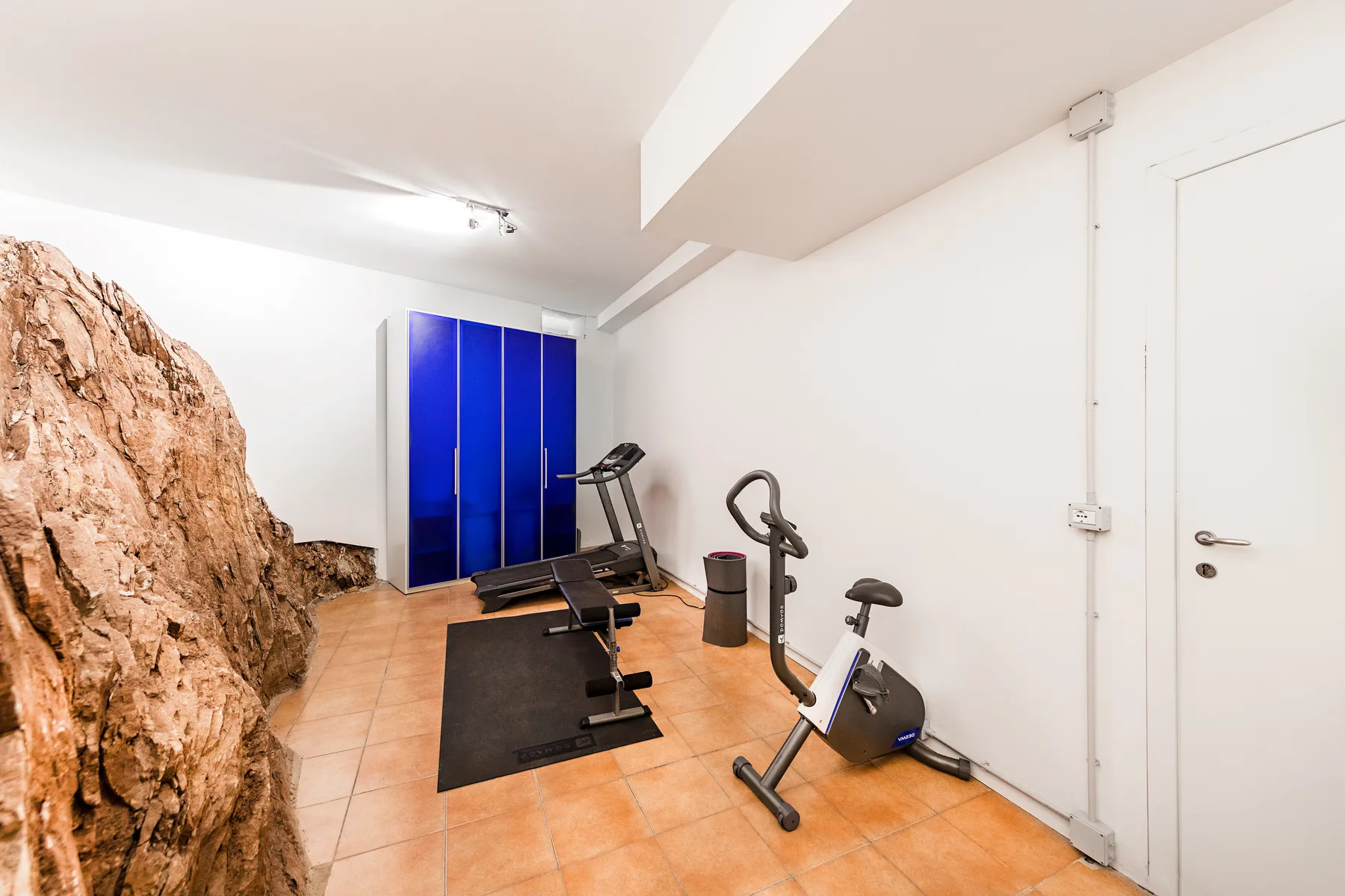Un pequeño gimnasio con máquinas de ejercicio, un armario azul y roca expuesta.