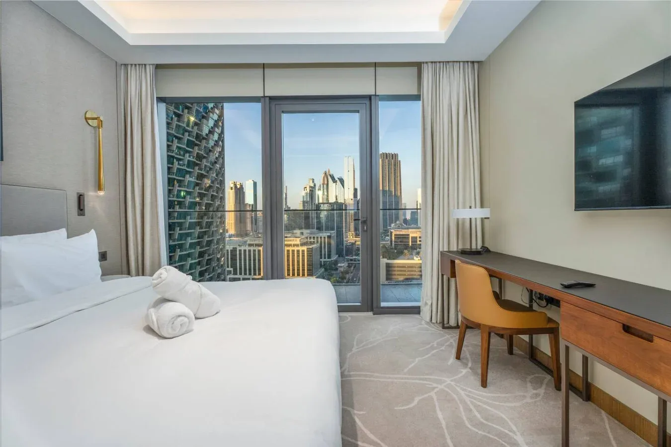 A bright bedroom with a bed, desk, and city views through the windows.