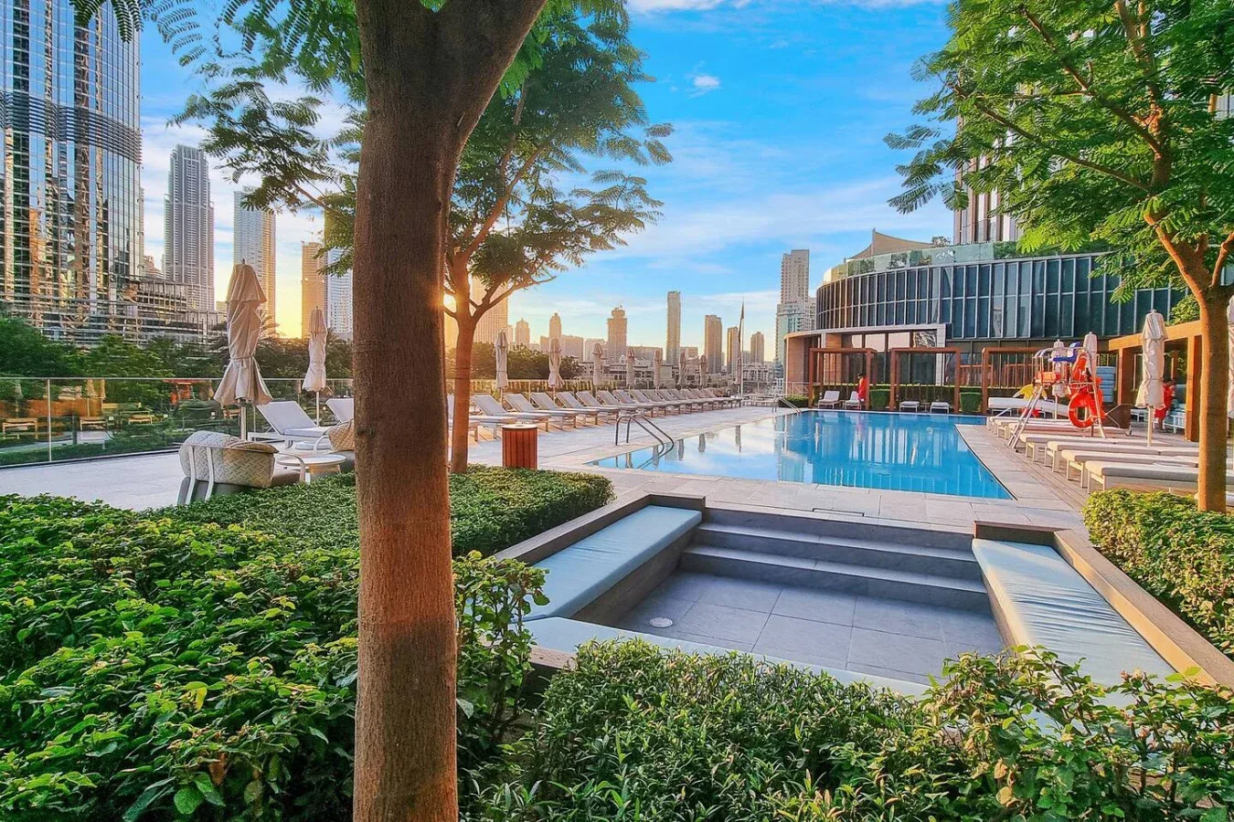 An outdoor swimming pool is surrounded by lounge seating, greenery, and tall city buildings.