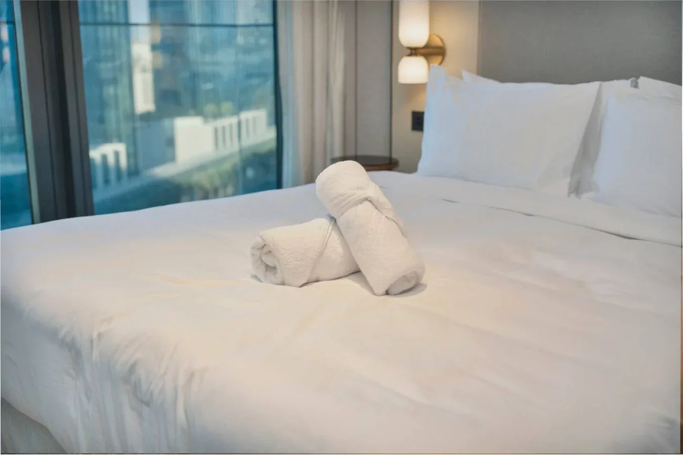 A neatly made bed with white linens and two rolled towels sits beside a large window.