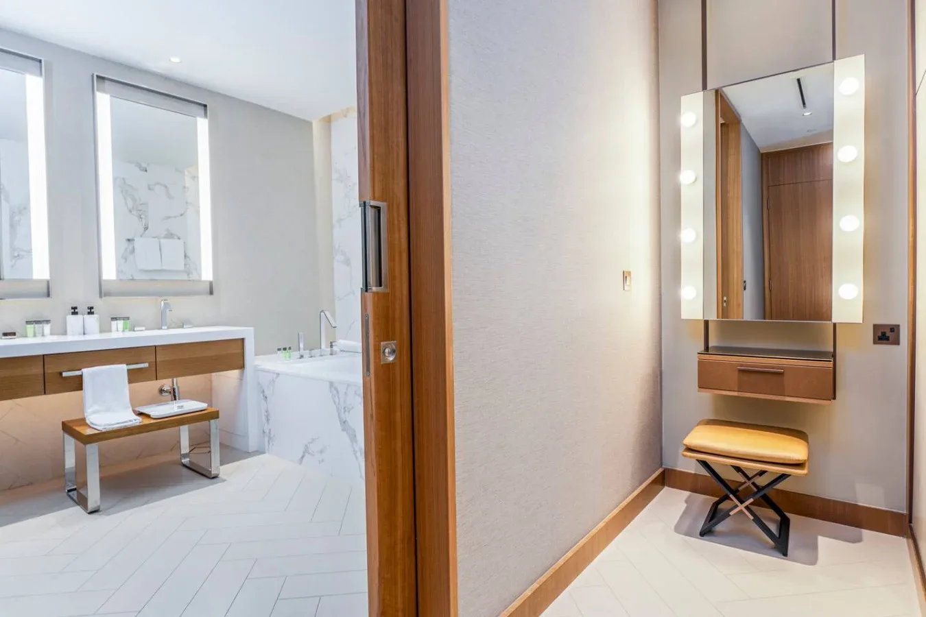 A bathroom vanity and bathtub sit beside a lit dressing mirror and bench.