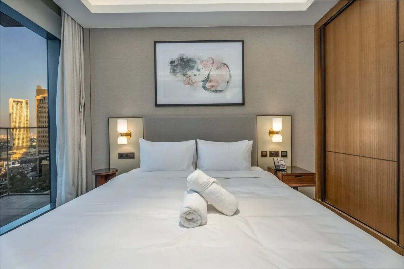 A bedroom with a neatly made bed, bedside tables, and a large window overlooking city buildings.