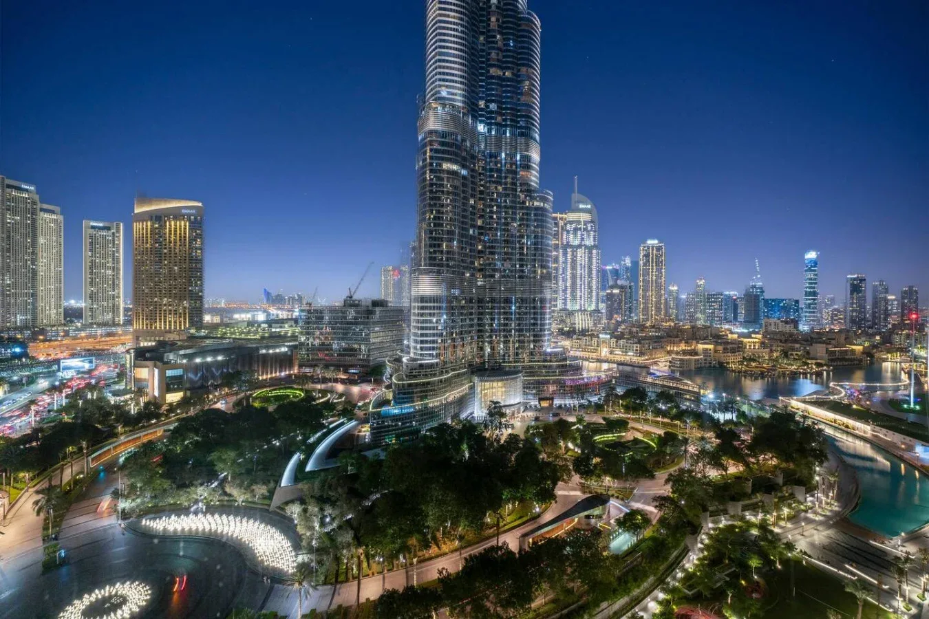 A nighttime city skyline with illuminated towers, landscaped paths, and a curving waterway.